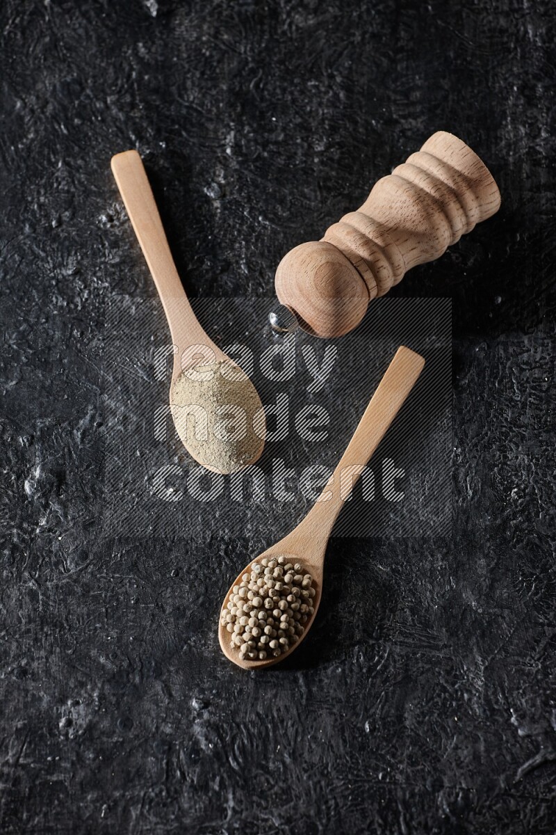 2 wooden spoons one full of white pepper powder and the other with pepper beads and a wooden pepper mill on textured black flooring