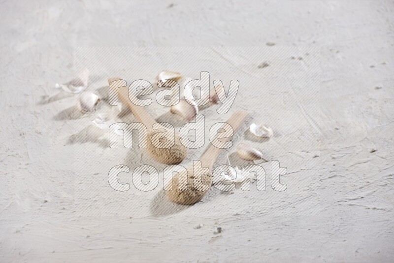 Two wooden spoons filled with garlic powder, surrounded by peeled garlic cloves and their skins on a textured white flooring