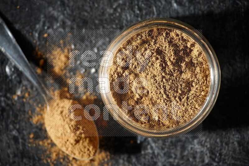 A glass jar and a metal spoon full of allspice powder on a textured black flooring