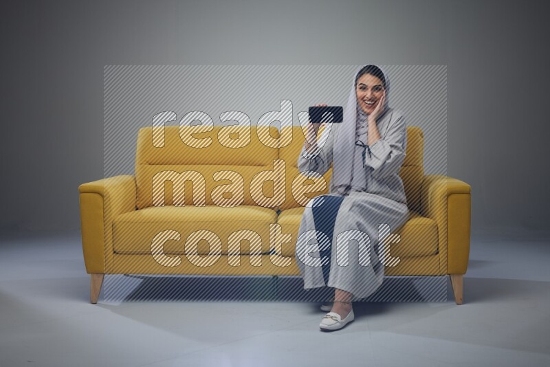 A Saudi woman wearing a light gray Abaya and head scarf setting on a yellow sofa and holding her phone while showing it's screen eye level on a grey background