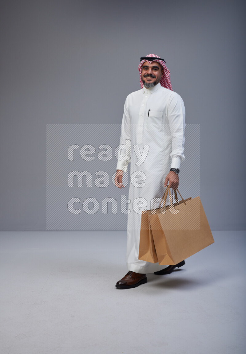 Saudi man Wearing Thob and red Shomag standing holding shopping bag on Gray background