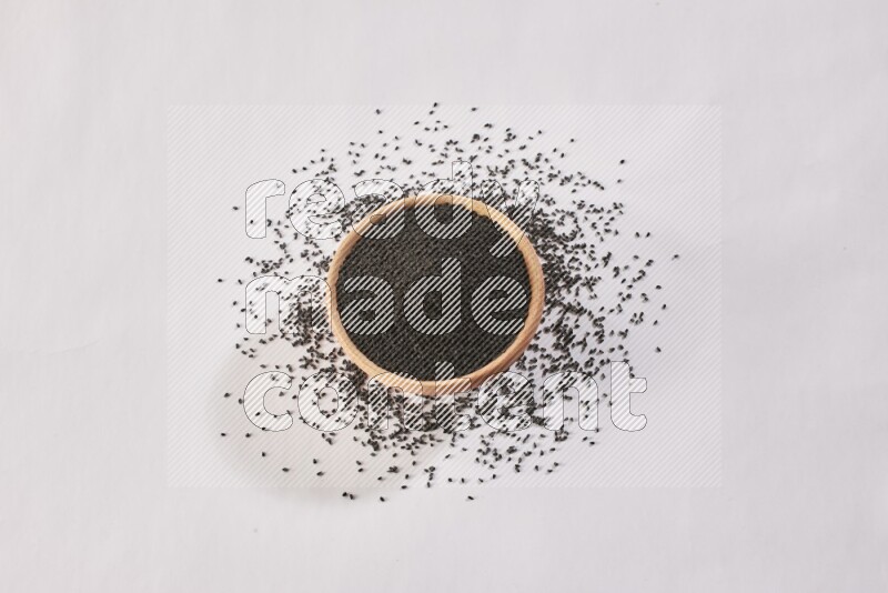 A wooden bowl full of black seeds and more seeds spread on a white flooring