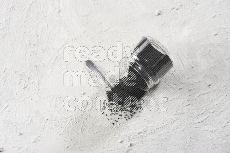 A glass jar and a metal spoon full of black seeds and the jar flipped and seeds spread on a textured white flooring