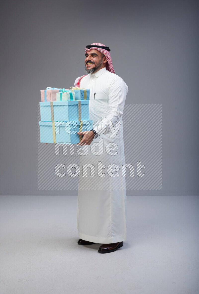 Saudi man Wearing Thob and red Shomag standing holding gift box on Gray background