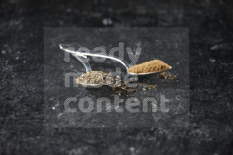 2 metal spoons full of cumin seeds and cumin powder on a textured black flooring