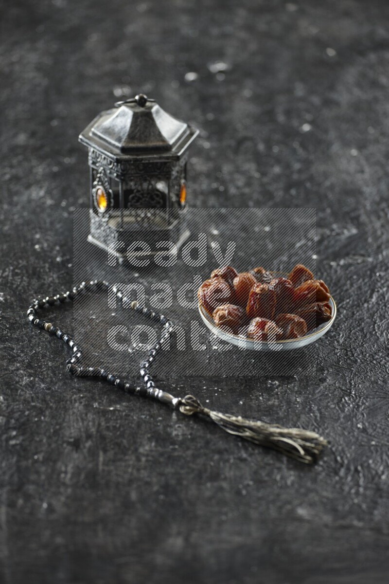 A silver lantern with different drinks, dates, nuts, prayer beads and quran on textured black background