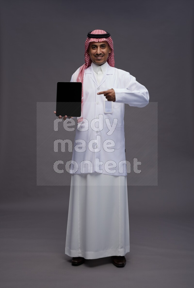 Saudi man wearing thob with lab coat and shomag with pocket employee badge standing showing tablet to camera on gray background
