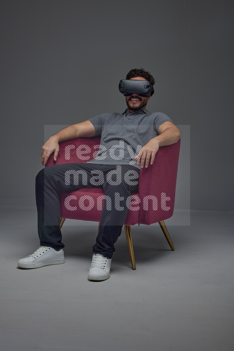 A man wearing casual and wearing VR while sitting on a burgundy chair eye level on a gray background