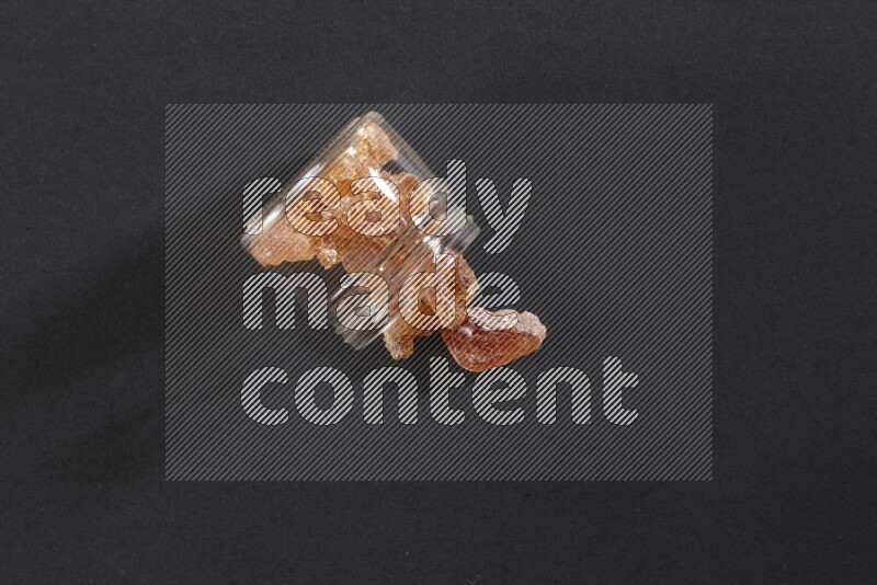 وعاء زجاجي ممتلئ بقطع الصمغ العربي والوعاء مقلوب وبعض القطع متناثرة خارجه علي خلفية سوداء