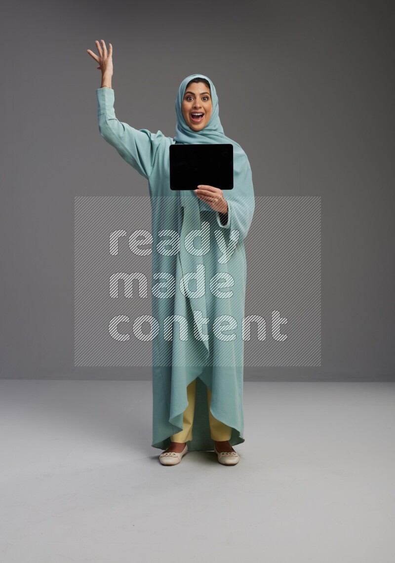 Saudi Woman wearing Abaya standing showing tablet to camera on Gray background