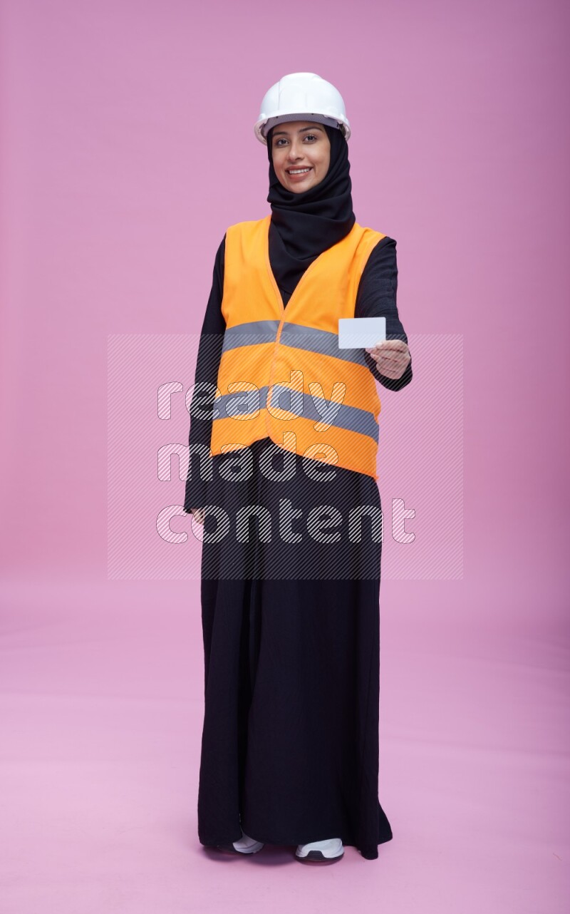 Saudi woman wearing Abaya with engineer vest and helmet standing holding ATM card on pink background