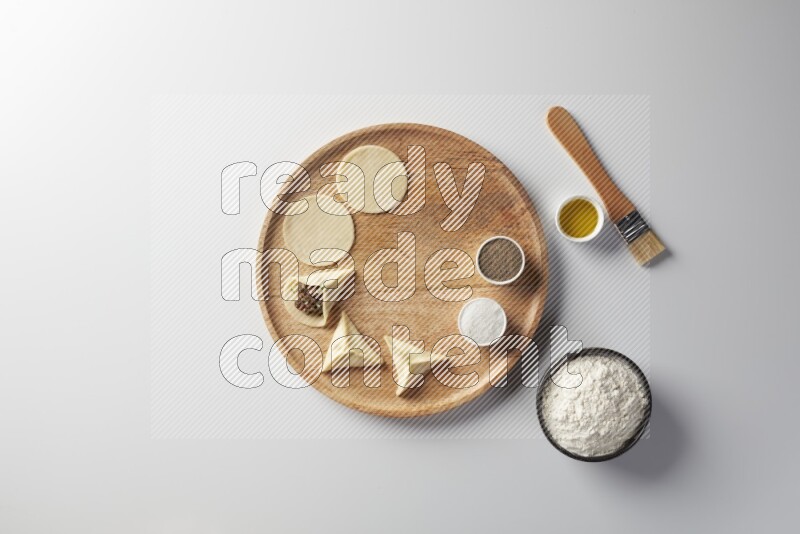 two closed sambosas and one open sambosa filled with meat while flour, salt, black pepper and oil with oil brush aside in a wooden dish on a white background