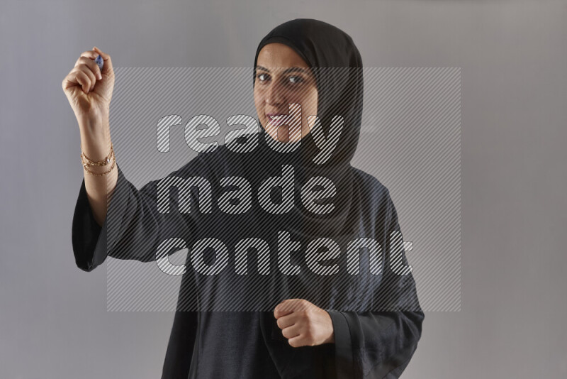 Woman in black abaya and hijab holding a marker pen to write on a transparent board with different reactions and expressions on grey background