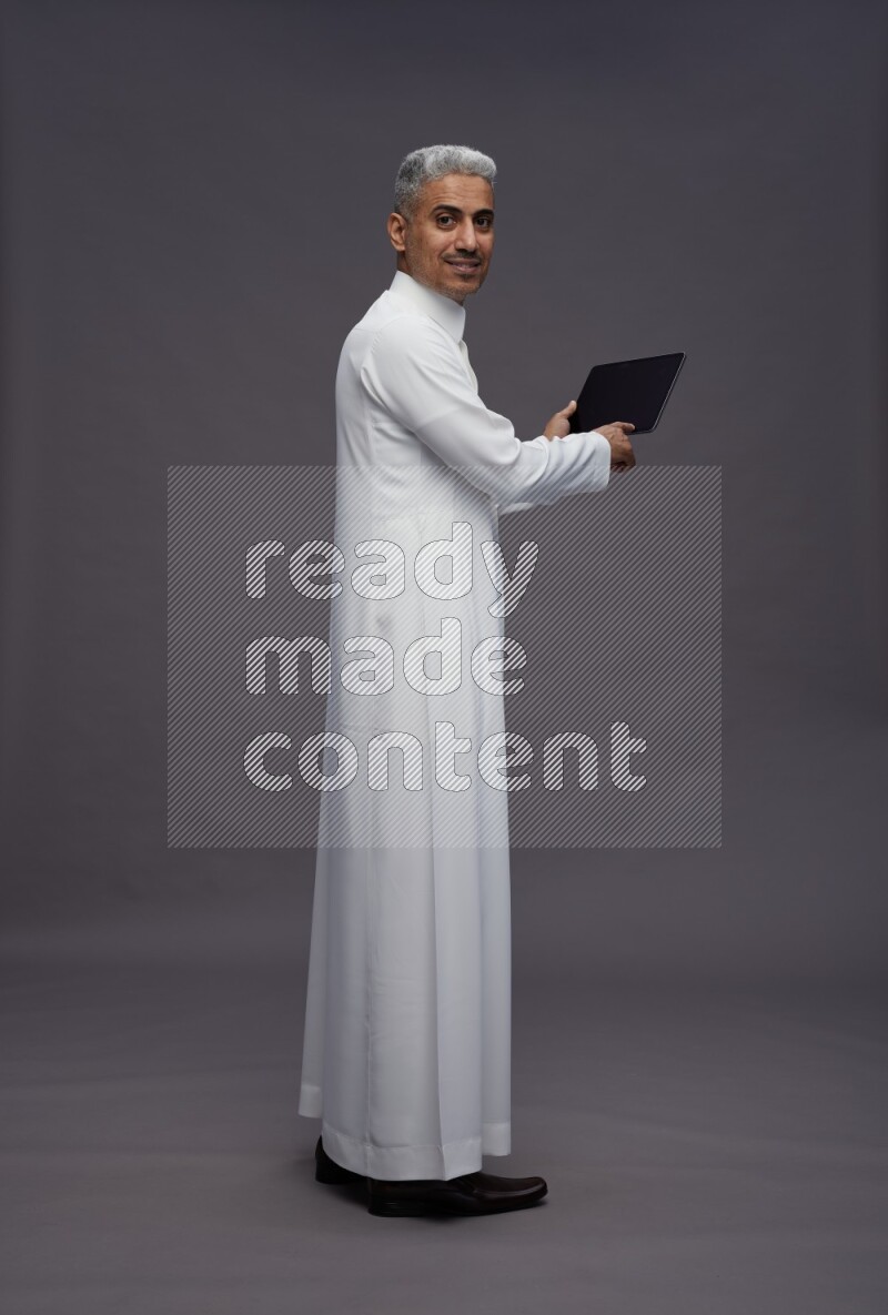 Saudi man wearing thob standing working on tablet on gray background