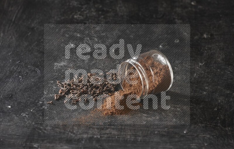 A flipped glass jar full of cloves powder with cloves spread on a textured black flooring