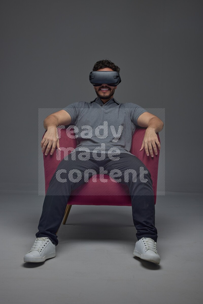 A man wearing casual and wearing VR while sitting on a burgundy chair eye level on a gray background