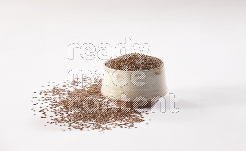 A pottery beige bowl full of flax seeds and more seeds spread on a white flooring