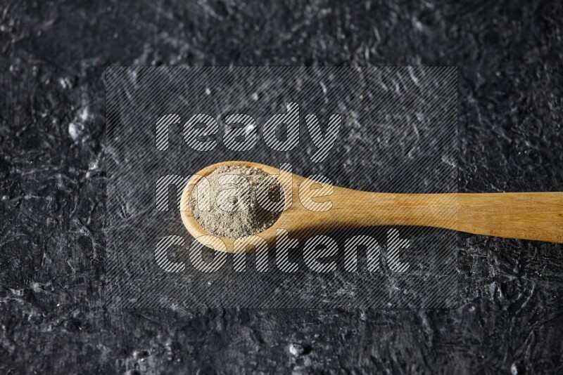A wooden spoon full of white pepper powder on textured black flooring