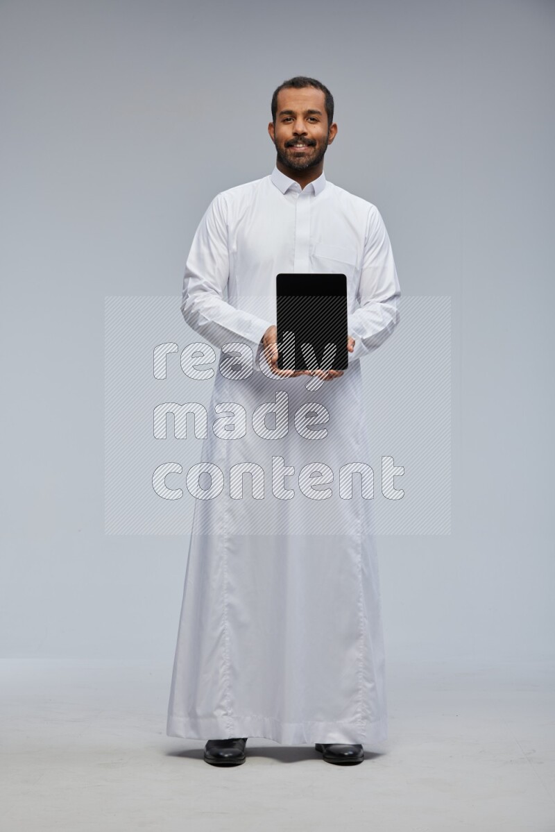 Saudi man wearing Thob standing showing tablet to camera on Gray background