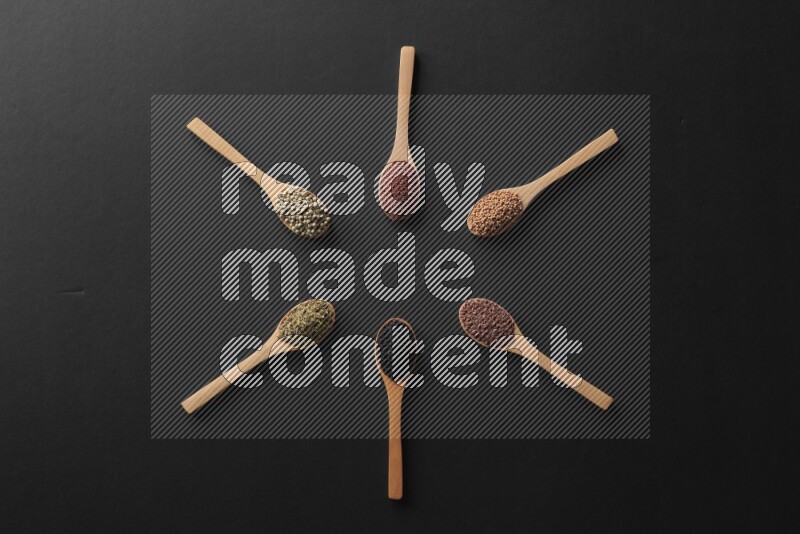 wooden spoons filled with white peppers, mustard seeds, black seeds, garden cress, cumin and flax on black flooring and shaped like a clock