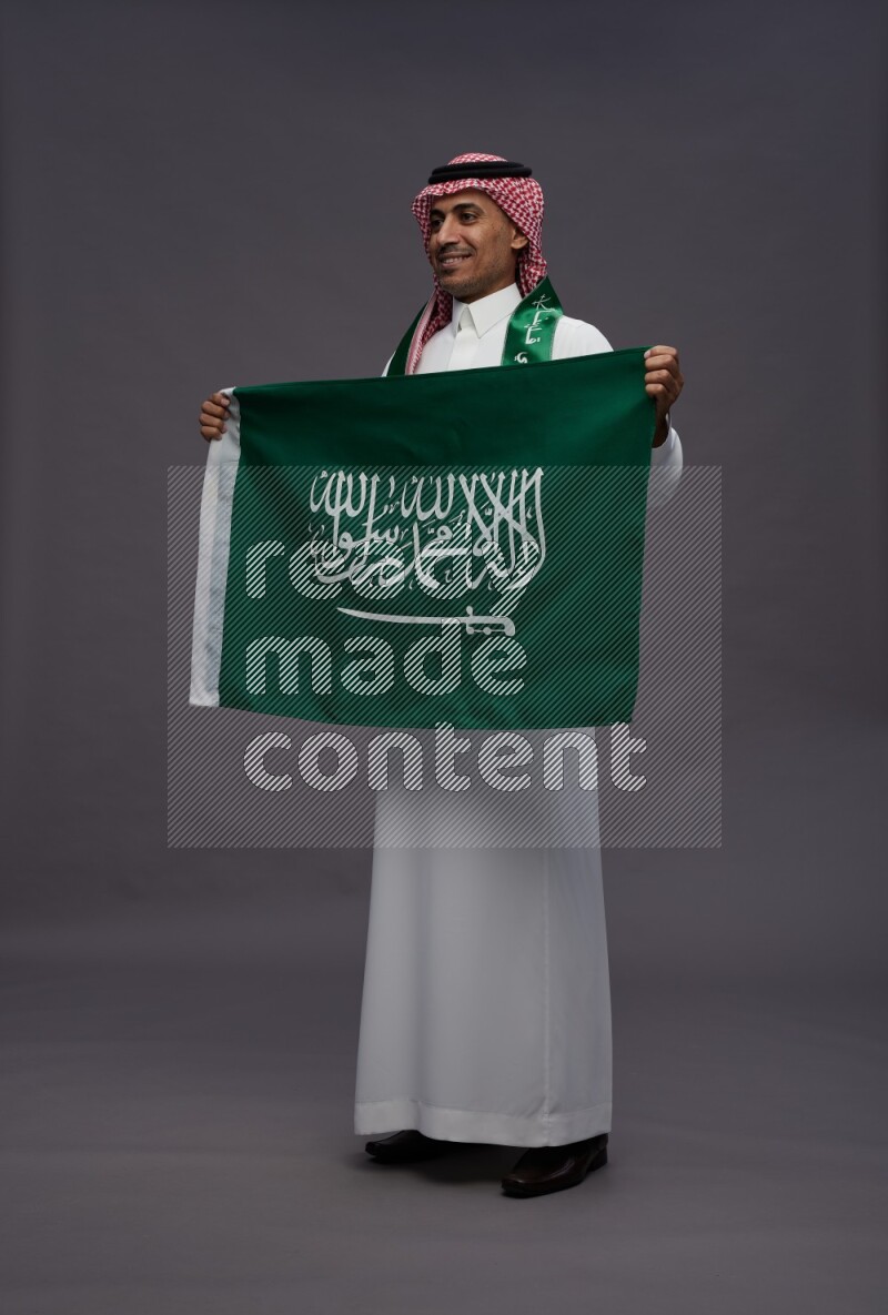 Saudi man wearing thob and shomag standing holding Saudi flag on gray background