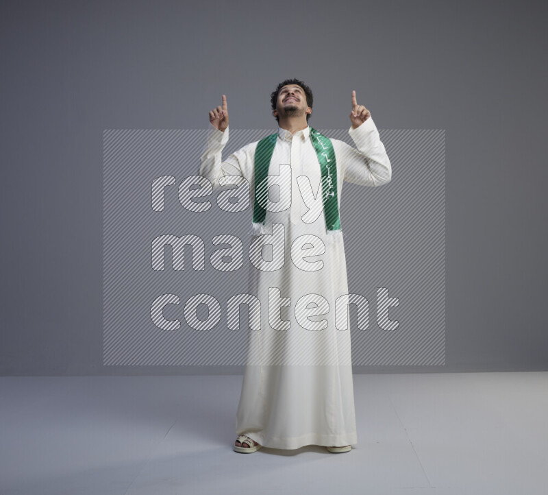 A Saudi man standing wearing thob and Saudi flag scarf on gray background