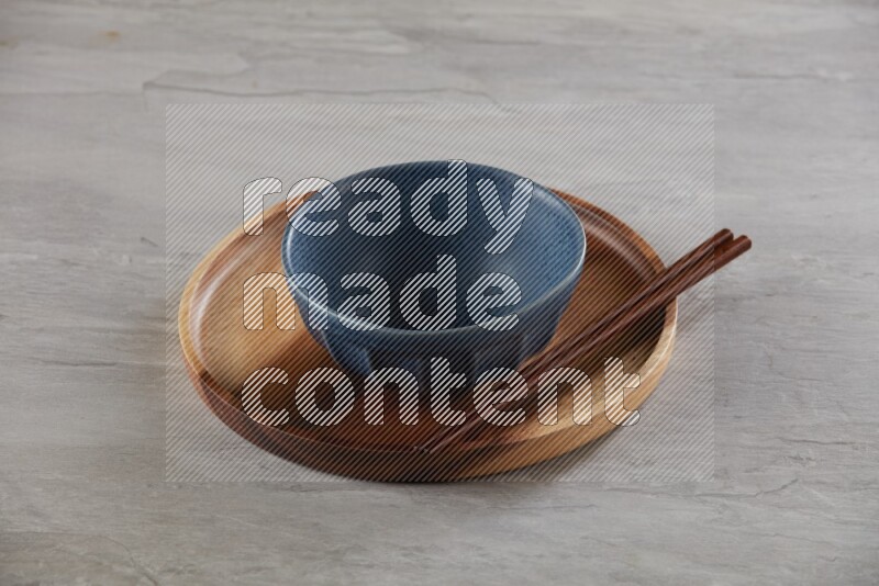 Blue pottery round bowl on top of brown wood round plate and wood chopsticks, on grey textured countertop
