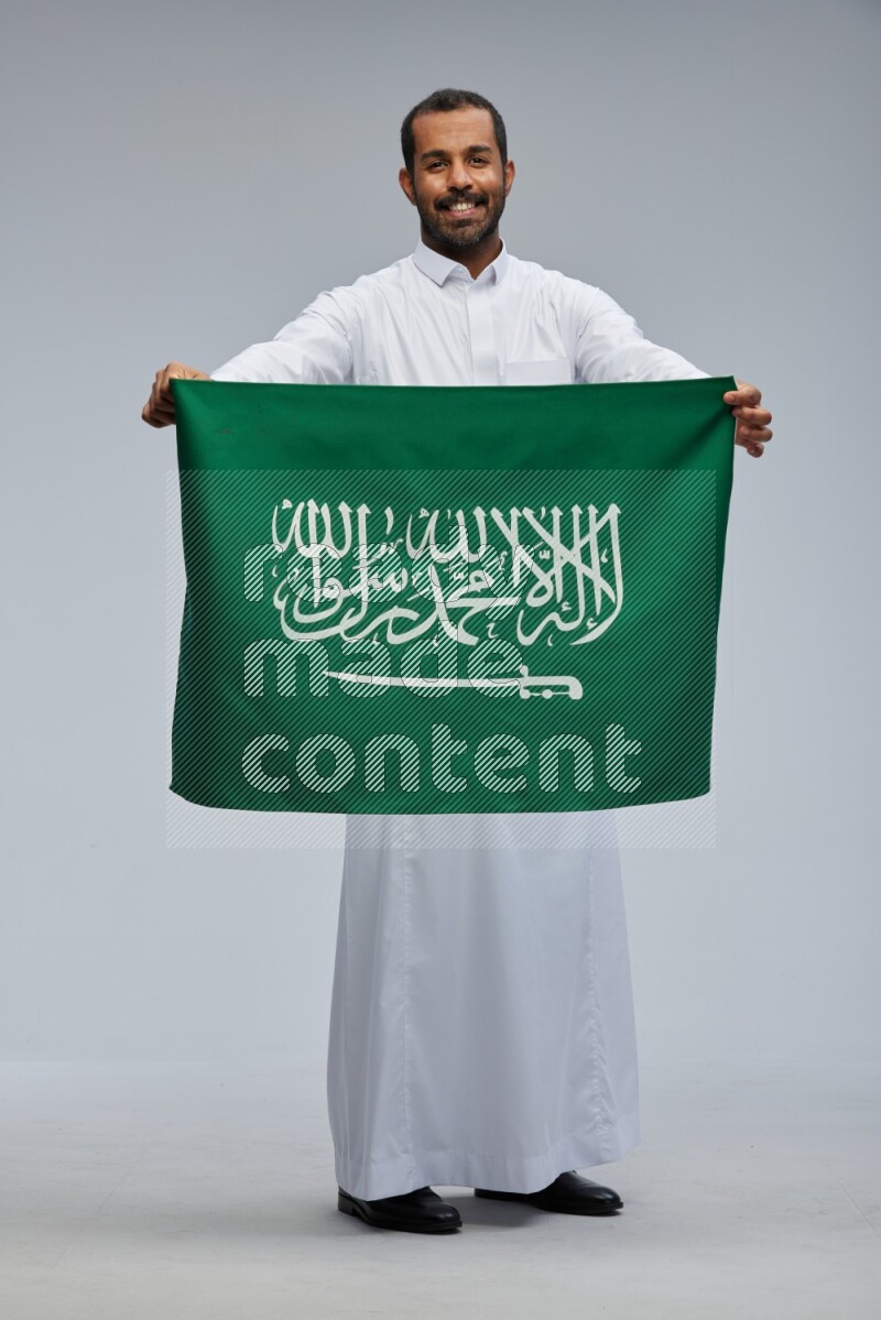 Saudi man wearing Thob standing holding Saudi flag on Gray background