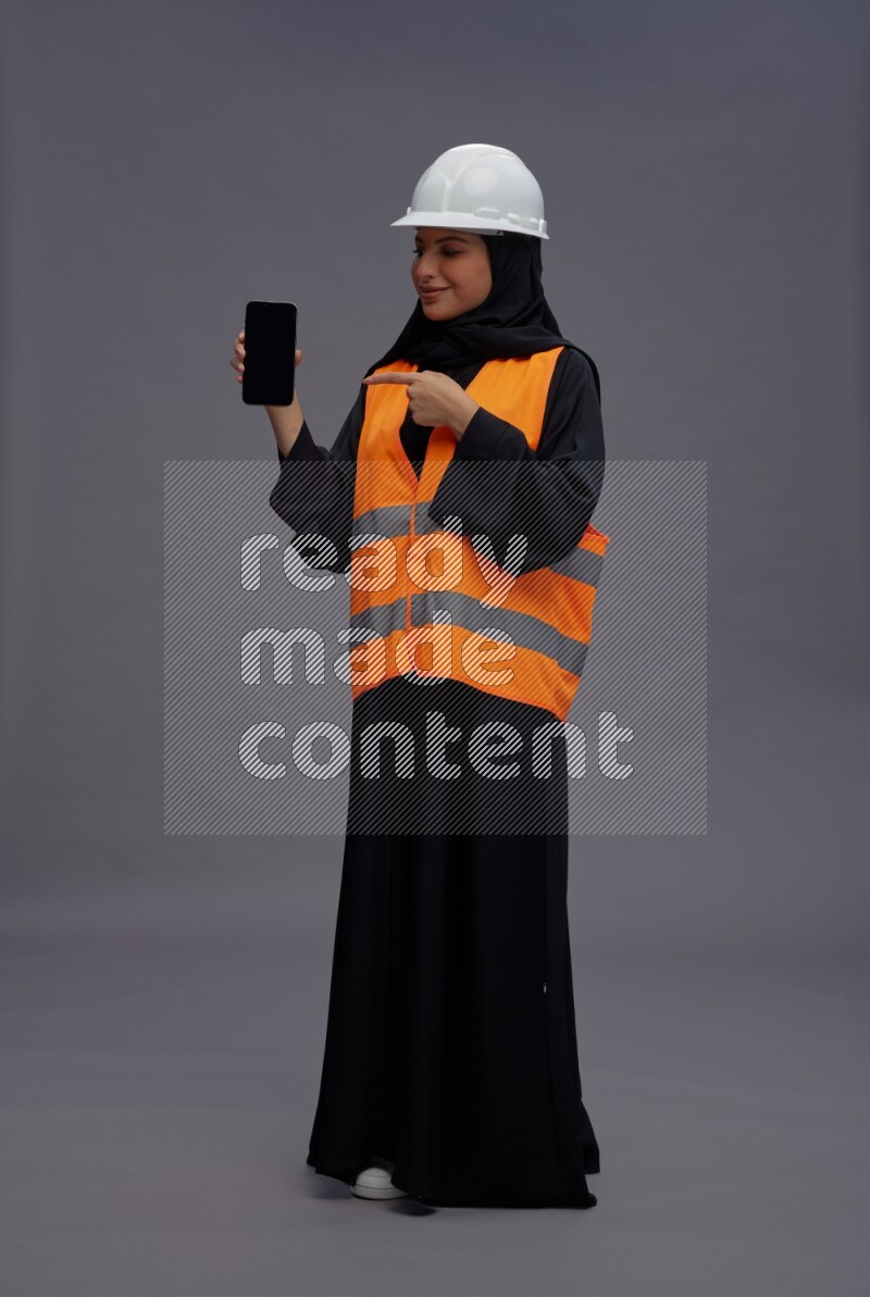 Saudi woman wearing Abaya with engineer vest standing showing phone to camera on gray background