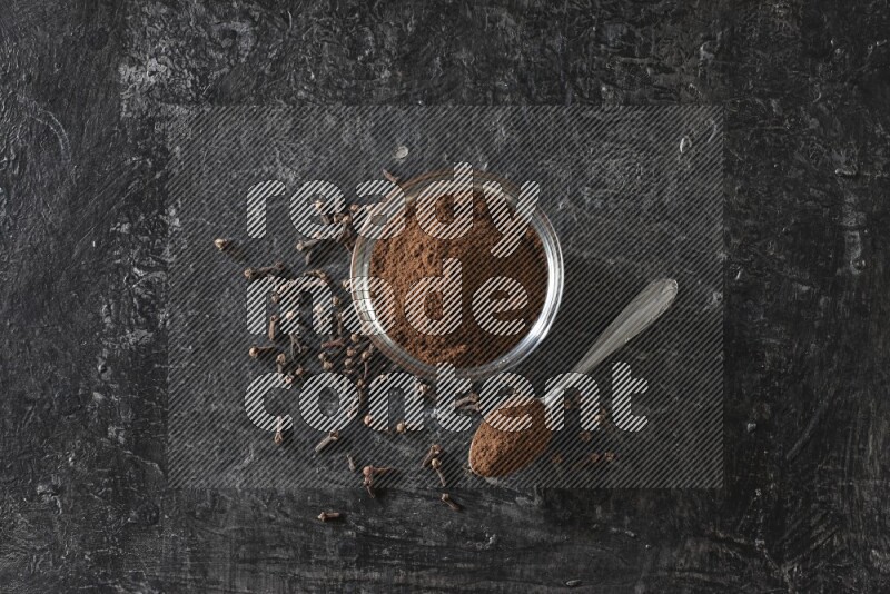 A glass bowl and a metal spoon full of cloves powder with gloves grains beside them on a textured black flooring