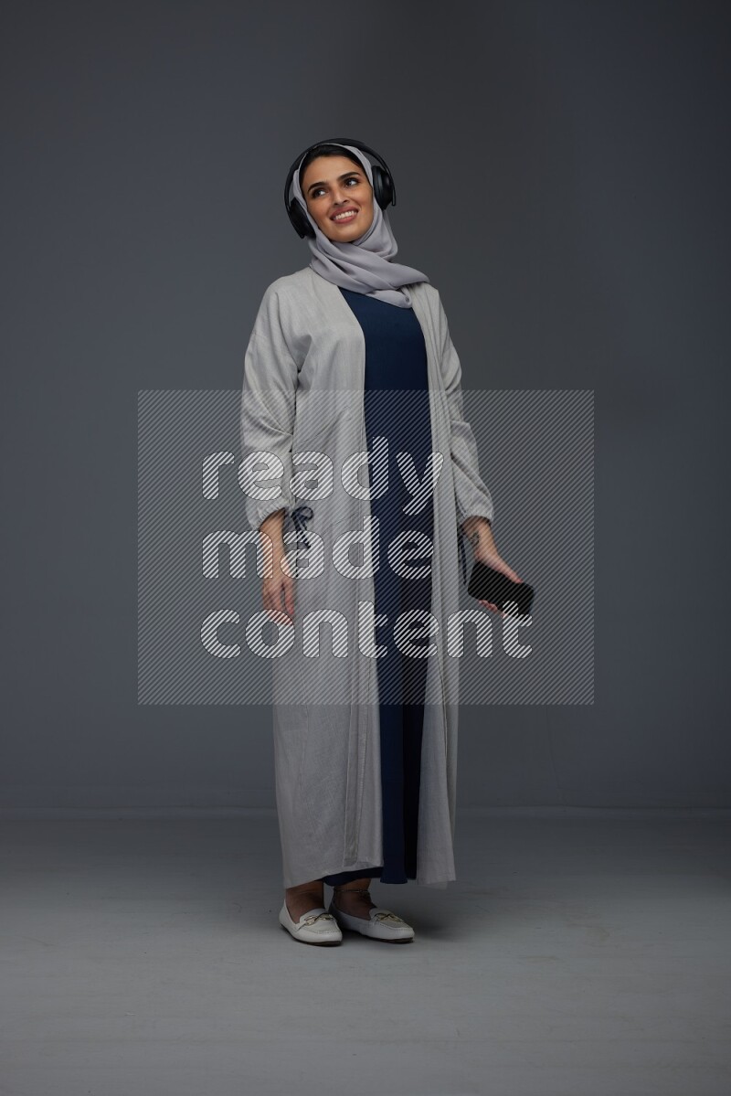 A Saudi woman wearing a light gray Abaya and head scarf standing and listening to music on a grey background