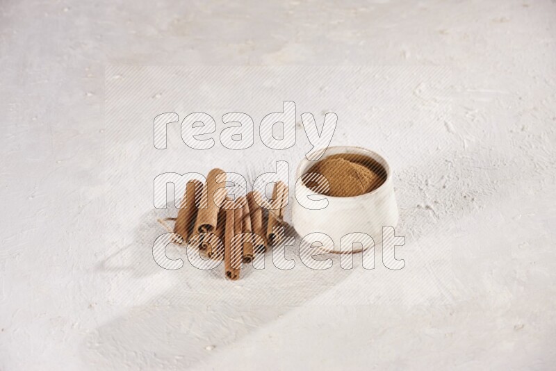 Cinnamon sticks stacked beside a beige bowl full of cinnamon powder on white background