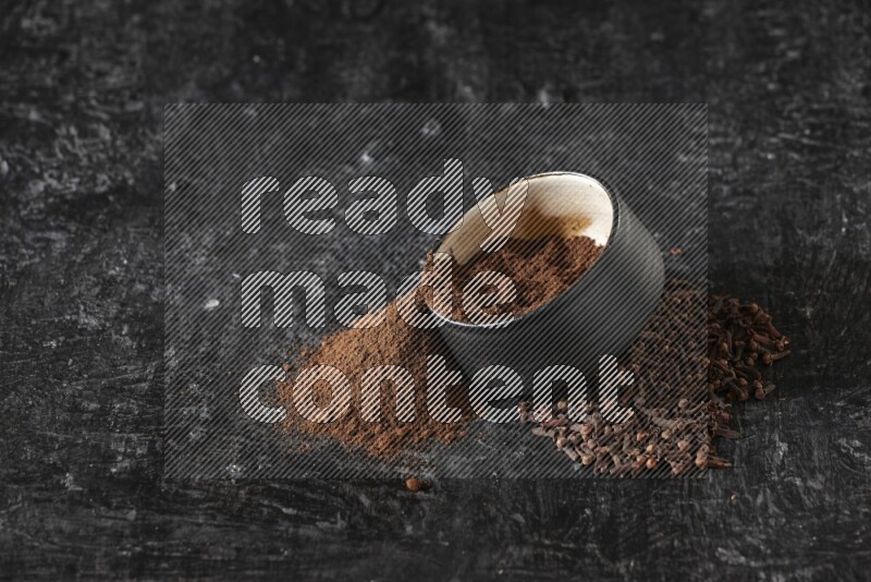 A black pottery bowl full of cloves powder spilled out of it with whole grains on a textured black flooring