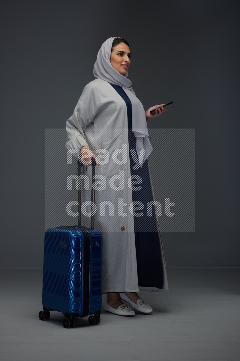 A Saudi woman wearing a light gray Abaya and head scarf standing holding a phone and being shocked  eye level on a grey background