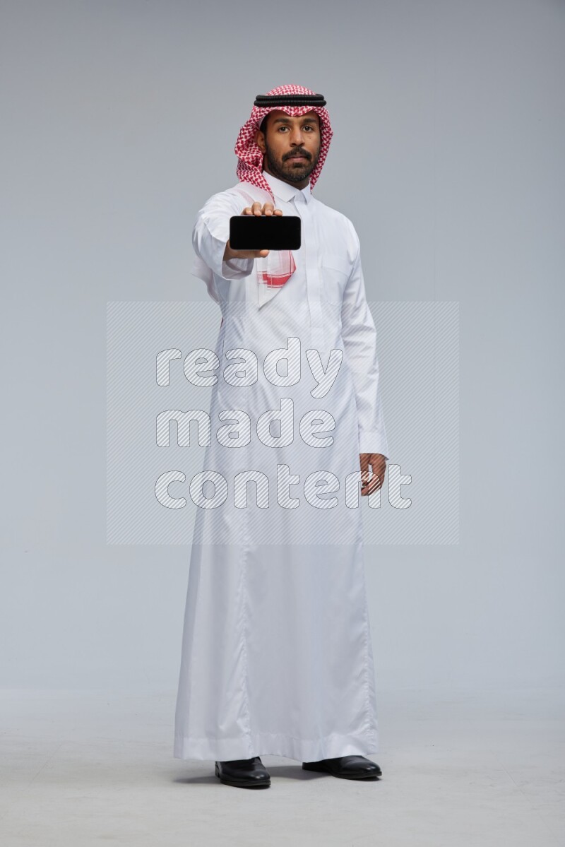 Saudi man Wearing Thob and shomag standing showing phone to camera on Gray background