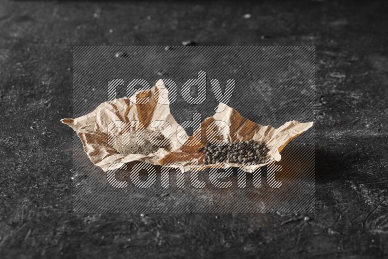 Black pepper and black pepper powder on 2 crumpled paper on a textured black flooring