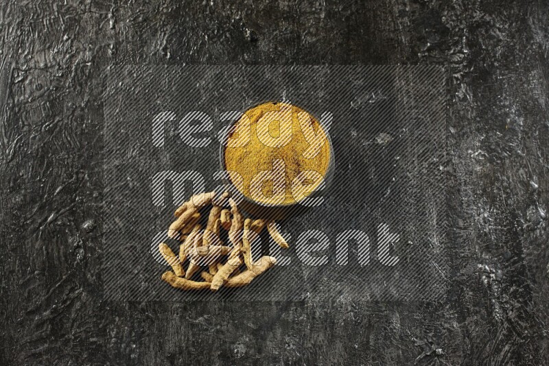 وعاء فخار أسود ممتلئ ببودرة الكركم وأصابع الكركم المجفف بجانبه على خلفية سوداء