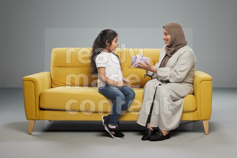 A girl sitting on a yellow sofa and giving a gift to her mother on gray background