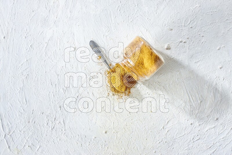 وعاء زجاجي مقلوب وملعقة معدنية ممتلئان ببودرة الكركم والبودرة تناثرت منه على خلفية بيضاء