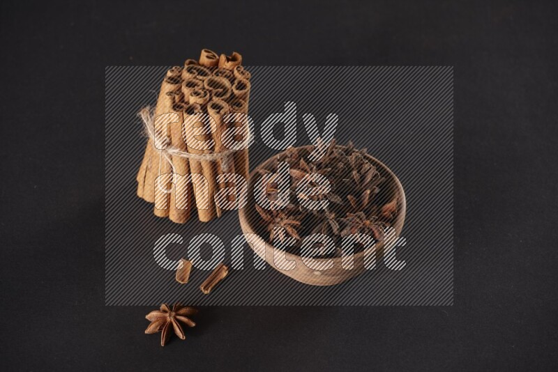A stacked and bounded cinnamon sticks and a wooden bowl full of star anise on a black background