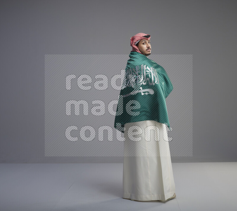 A Saudi man standing wearing thob and red shomag wrapping big saudi flag on gray background