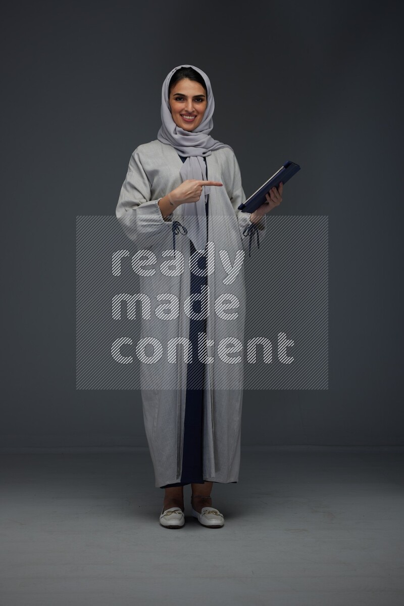 A Saudi woman wearing a light gray Abaya and head scarf standing holding a phone and pointing with the other hand eye level on a grey background