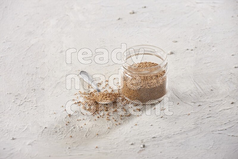 A glass jar and a metal spoon full of mustard seeds on a textured white flooring