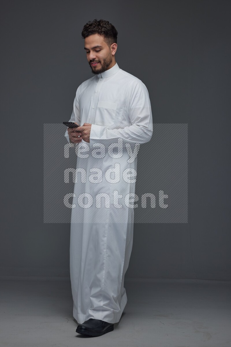 A Saudi man wearing Thobe standing and using his phone eye level on a gray background