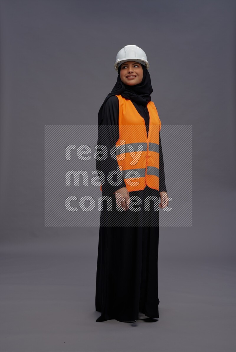 Saudi woman wearing Abaya with engineer vest standing interacting with the camera on gray background