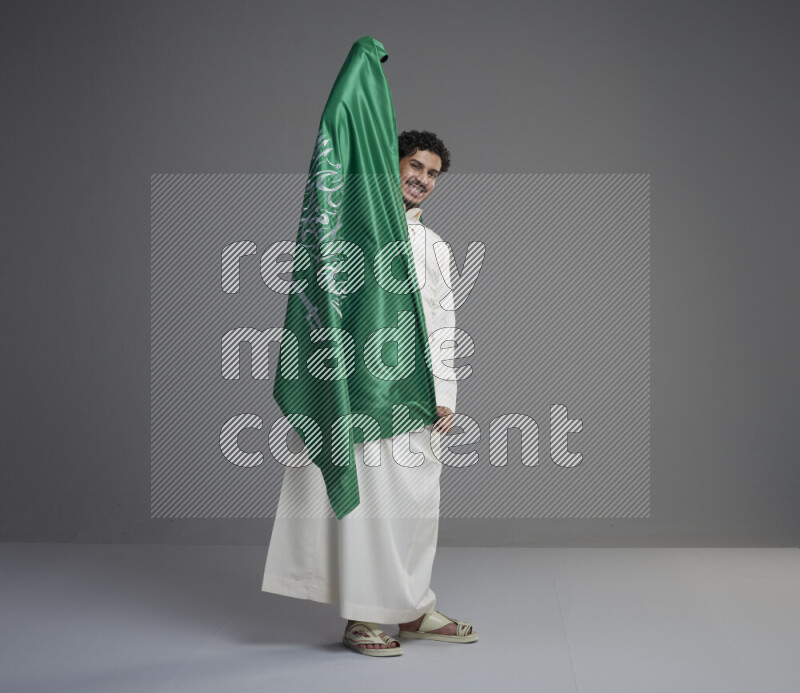 A Saudi man standing wearing thob holding big Saudi flag on gray background