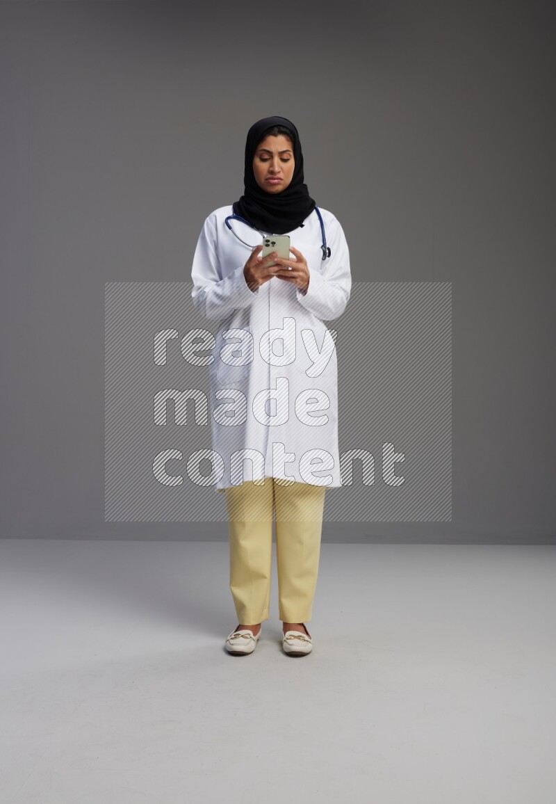 Saudi woman wearing lab coat with stethoscope standing texting on phone on Gray background