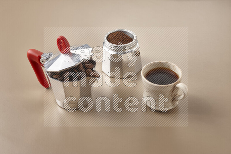 A moka pot with red handle surrounded by roasted coffee beans on beige background