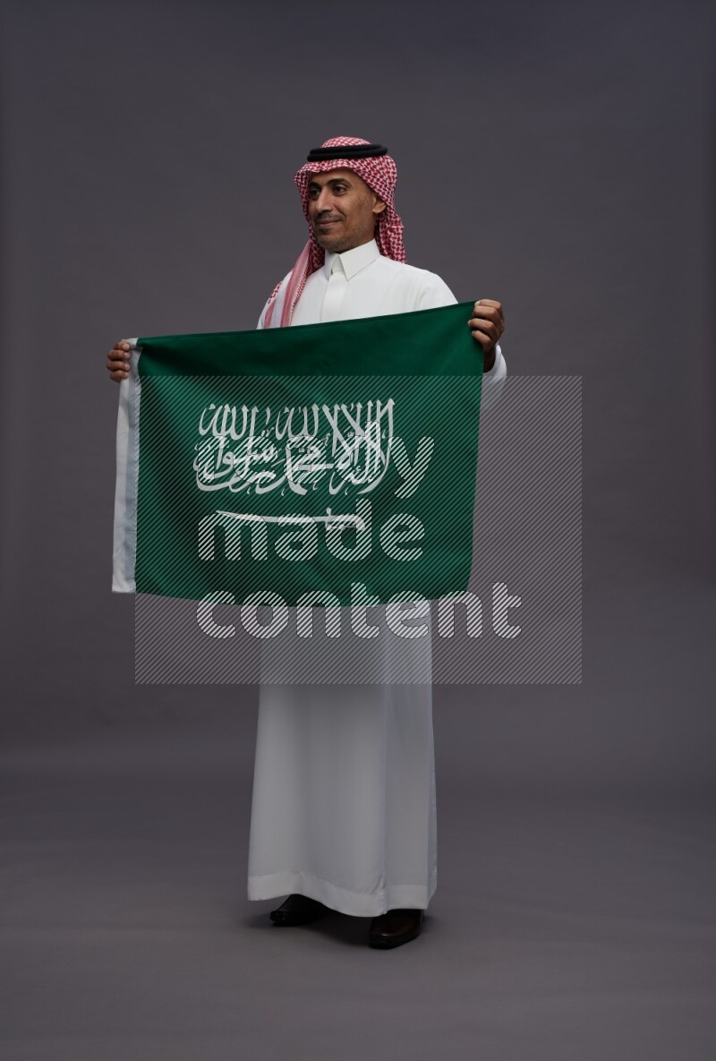 Saudi man wearing thob and shomag standing holding Saudi flag on gray background