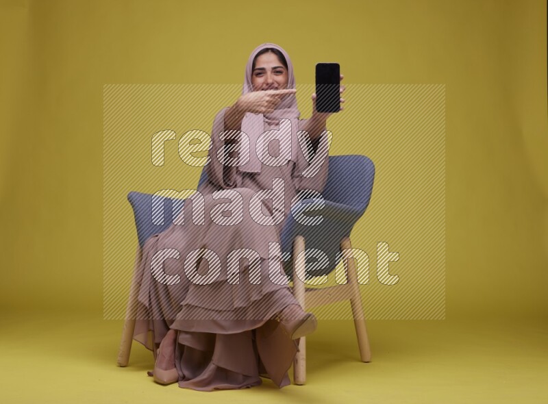A woman Showing Her Phone Screen Setting  on a Yellow Background wearing Brown Abaya with Hijab